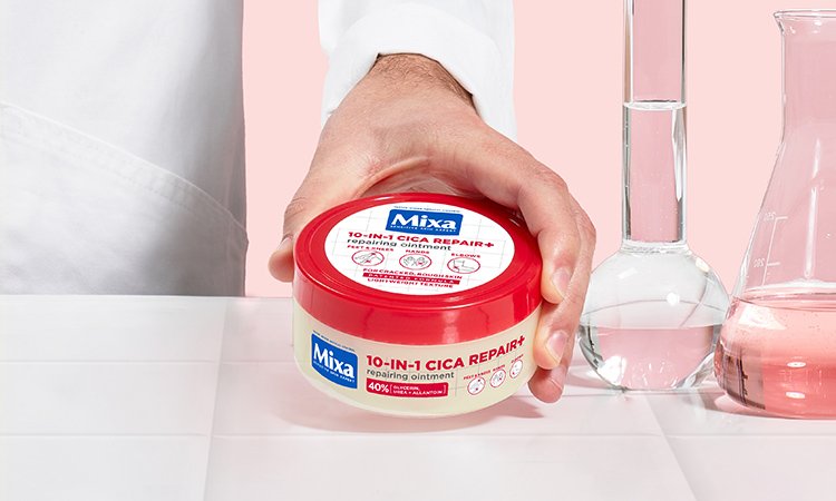 Produktabbildungen von MIXA Eine Person in weißem Kittel stellt die 10-in-1 Cica Repair+ Creme von Mixa auf einer weißen Fläche neben drei Reagenzgläsern mit rosafarbener und durchsichtigen Flüssigkeiten ab