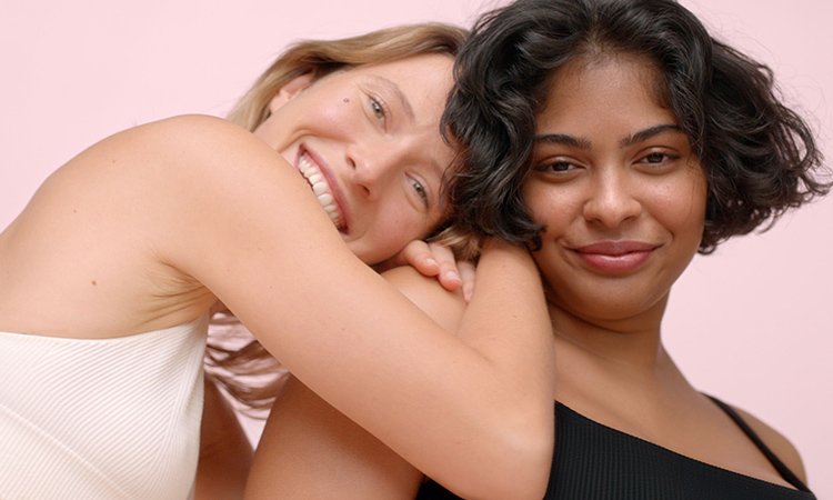 Eine lächelnde Frau lehnt sich mit beiden Armen auf die Schultern einer zweiten Frau Eine lächelnde Frau lehnt sich mit beiden Armen auf die Schultern einer zweiten Frau