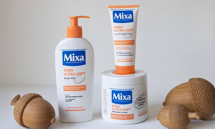 Die drei Mixa Produkte der Serie Shea Ultra Soft, die eine Body Milk, eine Hand- und Nagelcreme und eine nährenden Creme umfasst, stehen neben drei Eicheln vor einem hellen Hintergrund