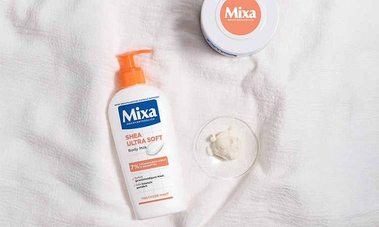 Die Mixa Shea Ultra Soft liegt auf einem weißen Tuch neben einer durchsichtigen Schale mit einem Klecks Sheabutter und einer Allzweckcreme von Mixa