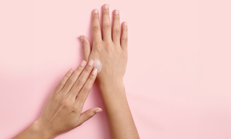 Nahaufnahme von zwei Händen einer Person die ihre Hände flach auf einer geraden Oberfläche liegen hat. Mit der linken Hand cremt sie ihre rechte Hand ein.
