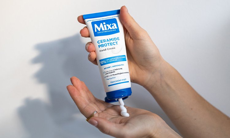 Zwei Hände einer Person mit einem Ring am linken Zeigefinger halten eine Handcreme, während sie die Creme auf die linke Hand aufträgt.
