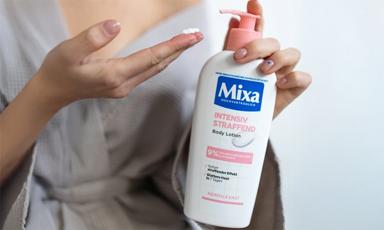 Eine Frau hält mit ihrer linken Hand die MIXA intensiv straffende Body Lotion und pumpt mit dem Spender einen Klecks Creme in ihre rechte Hand.