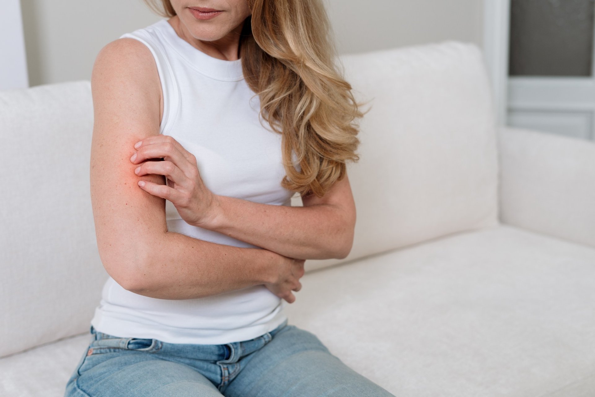 Frau sitzt auf einem Sofa und kratzt sich am Oberarm; sie trägt ein weißes ärmelloses Top und Jeans.