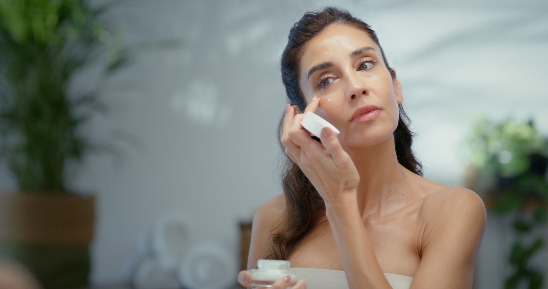 Frau trägt Gesichtspflege auf und tupft Creme unter das Auge; sie hält einen Cremetiegel in der Hand, im Hintergrund ein helles Badezimmer