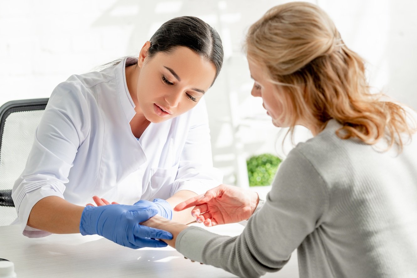Ärztin mit blauen Einmalhandschuhen untersucht in einer hellen Praxis das Handgelenk einer Patientin, die ihr den Arm über den Tisch reicht.