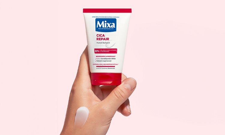 Mixa Creme wird in einer Hand gehalten vor rosa Hintergrund