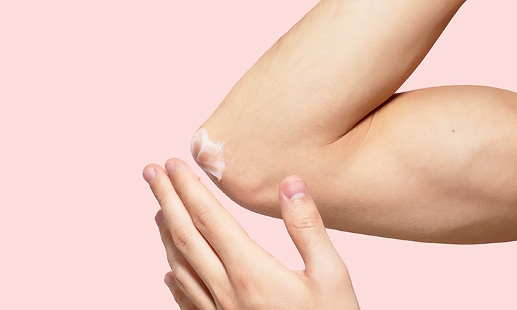 Eine Nahaufnahme zeigt einen Ellbogen, der von einer linken Hand mit Creme eingecremt wird.