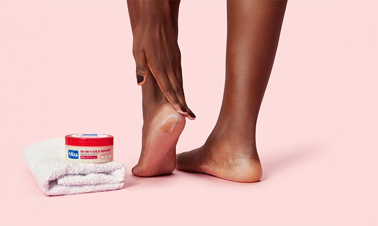 Eine Nahaufnahme zeigt ein Bein und einen Fuß. Eine linke Hand cremt die Ferse des linken Fußes ein. Auf dem Boden daneben liegt ein gefaltetes Handtuch mit einer Creme darauf.