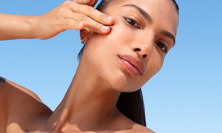 Eine Frau mit Zopf verteilt Sonnenschutzcreme auf ihrer Wange, im Hintergrund ein klarer blauer Himmel